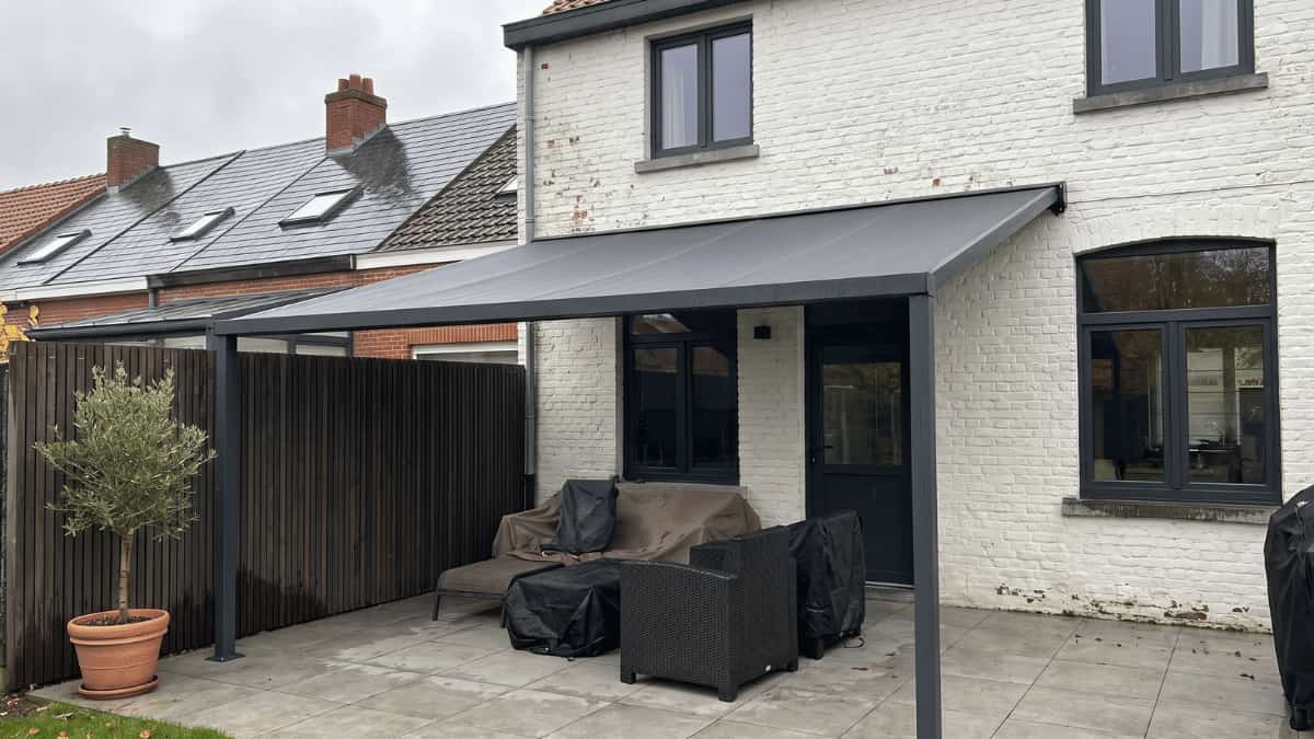 Een antraciet aluminium terrasoverkapping met gespannen grijs doek en twee verticale steunpalen staat tegen de witgeschilderde bakstenen gevel van een Belgische woning met donkere raamkozijnen terwijl eronder afgedekte tuinmeubelen en een rattan fauteuil op lichtgrijze betontegels staan naast een olijfboompje in een terracotta pot en een donker houten lamelscherm onder een bewolkte hemel.