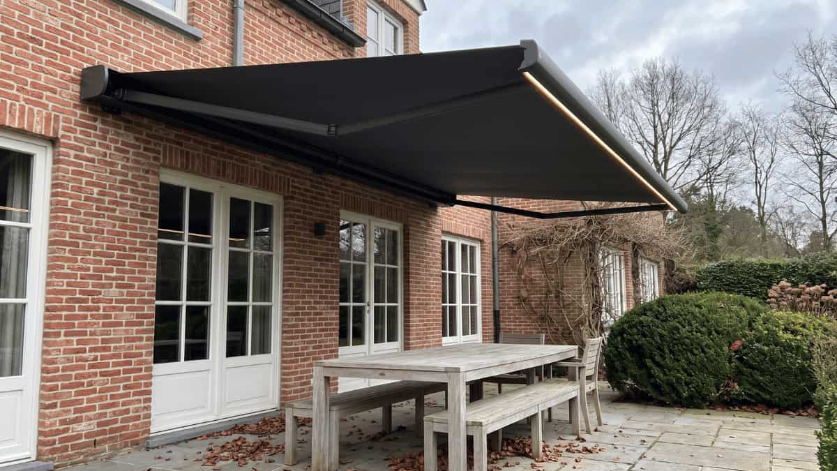 Een donker knikarmscherm met een warm verlichte ledstrip langs de voorrand hangt volledig uitgerold boven een verweerde houten tuintafel met bijpassende bank op een stenen terras tegen een Vlaamse bakstenen gevel met witgeschilderde openslaande deuren terwijl buxusstruiken en kale klimplanten de tuin omlijsten onder een bewolkte hemel.