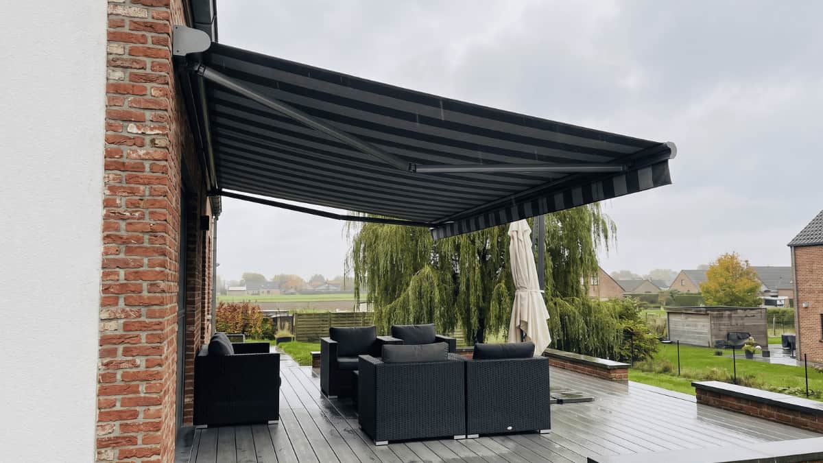 Een volledig uitgerold donkergrijs gestreept knikarmscherm beschaduwt een terras met grijze houten vlonders en antraciet rattan loungezetels tegen een gevel van rode baksteen en witte pleister onder een bewolkte Vlaamse hemel met een treurwilg en bakstenen woningen op de achtergrond.