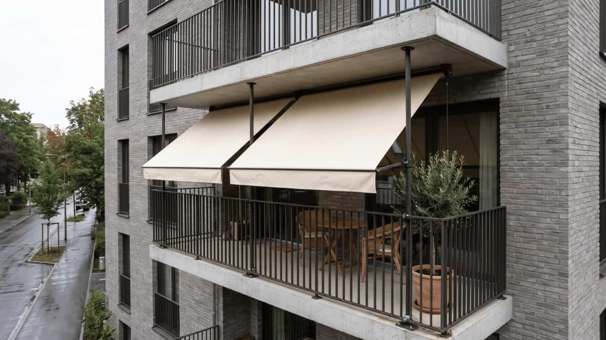 Twee crèmekleurige uitvalschermen aan donkere klemstangen beschaduwen een balkon met een houten tuinset en een olijfboompje in een terracotta pot op een modern grijs bakstenen appartementsgebouw op een bewolkte dag.