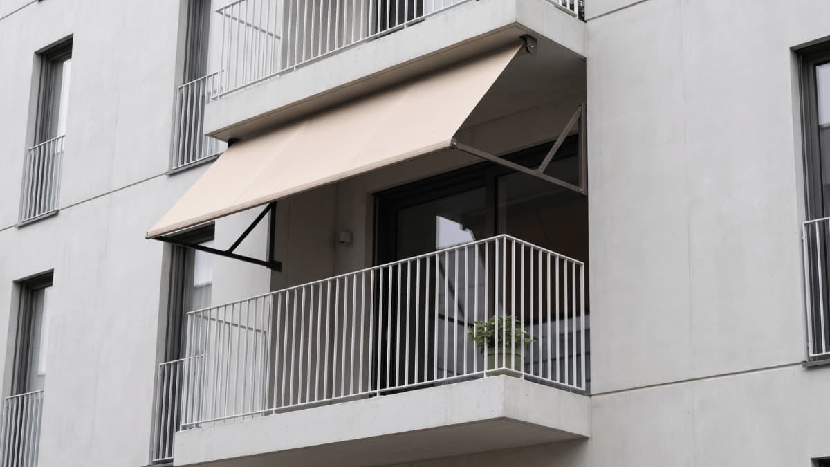 Een beige uitvalscherm met donkere metalen schaararmconstructie hangt schuin uitgeklapt boven een klein balkon met witte spijlenbalustrade en een groene potplant aan een strak grijs gepleisterd appartementsgebouw onder een bewolkte hemel.