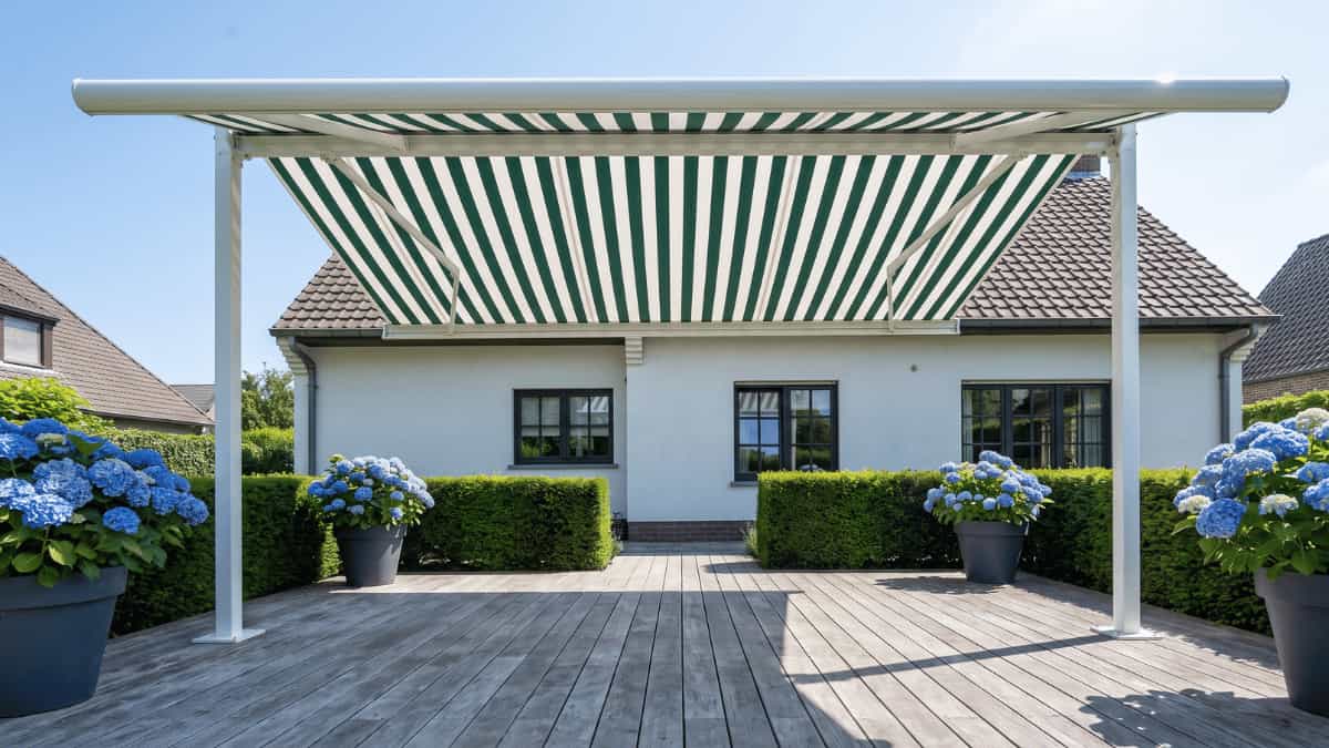Een vrijstaand zonnescherm met groenwit gestreept doek gespannen tussen twee witte aluminium palen overspant een grijs verweerd houten terras voor een witgepleisterde woning met donkere raamkozijnen en bruine dakpannen terwijl grote donkergrijze potten met felblauwe hortensia's aan weerszijden van de constructie staan opgesteld tegen een achtergrond van strak geschoren groene hagen en een heldere zomerlucht.