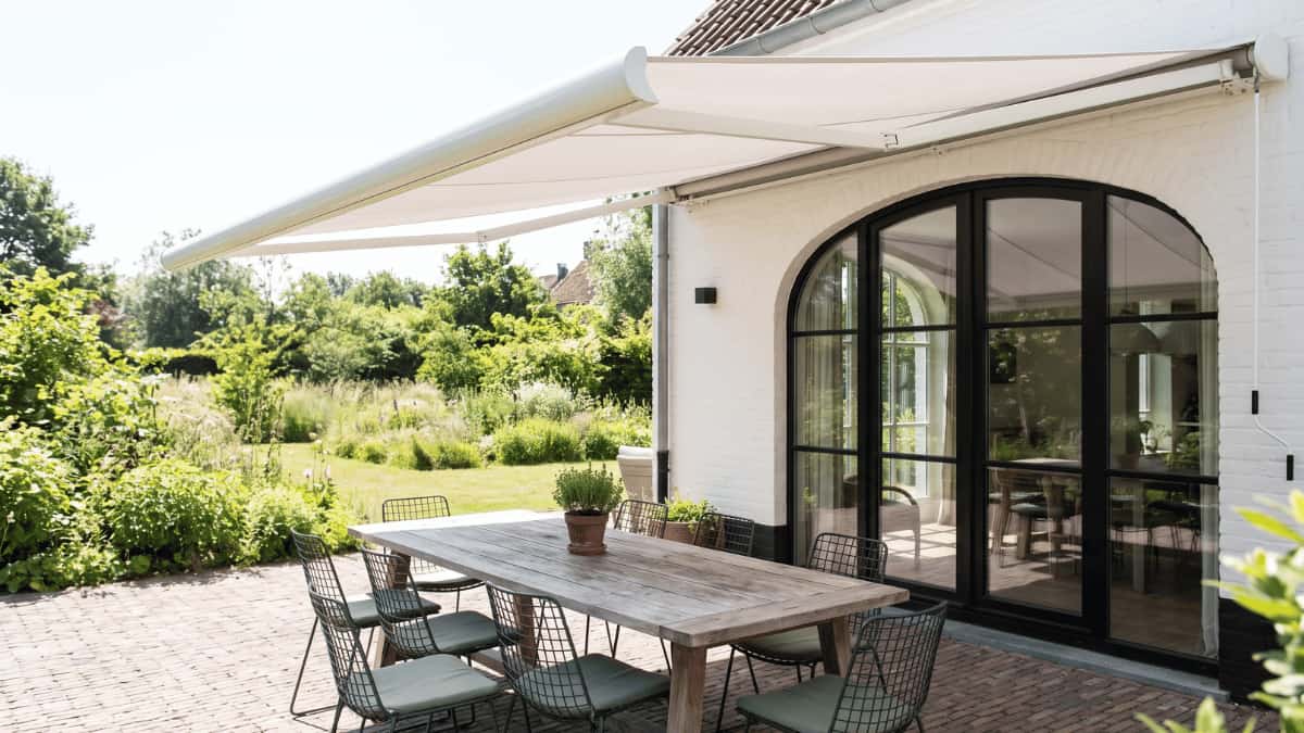 Een volledig uitgerold wit knikarmscherm overschaduwt een verweerde houten eettafel met draadstoelen en groene kussens op een klinkerterras tegen een witgeschilderde gevel met een groot boogvormig zwart omlijst glasraam en een weelderige groene tuin op de achtergrond.